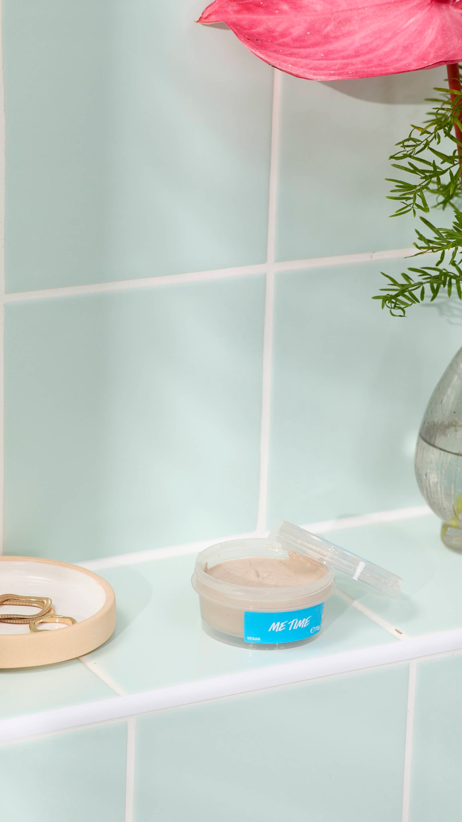 A small, clear pot of 'ME TIME' fresh face mask sits open on a pale green tiled ledge. A dab of the creamy, beige-coloured mask is on the lid. Beside it is a small ceramic dish with gold jewellery and a vase with a pink flower.