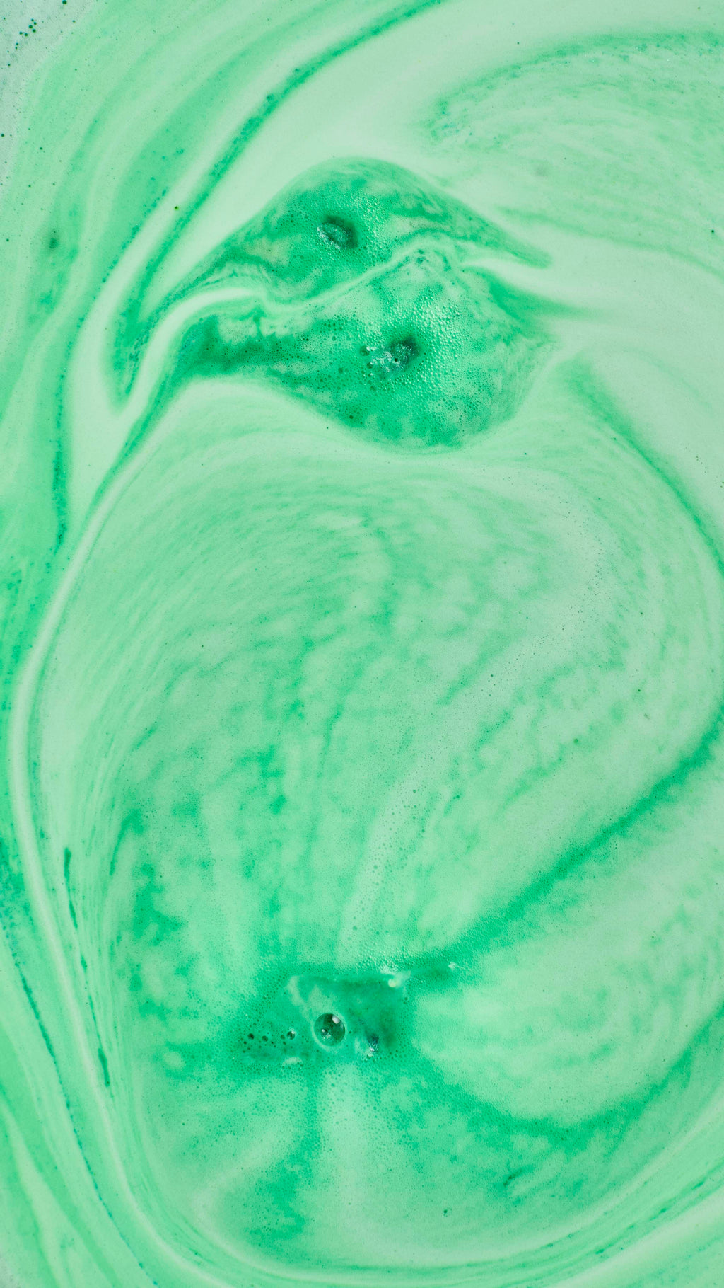 An abstract aerial shot of bath bomb foam swirling in water, creating marbled, wavy patterns of vibrant lime green and pale mint green. A few small clusters of soapy bubbles are scattered across the frothy surface.