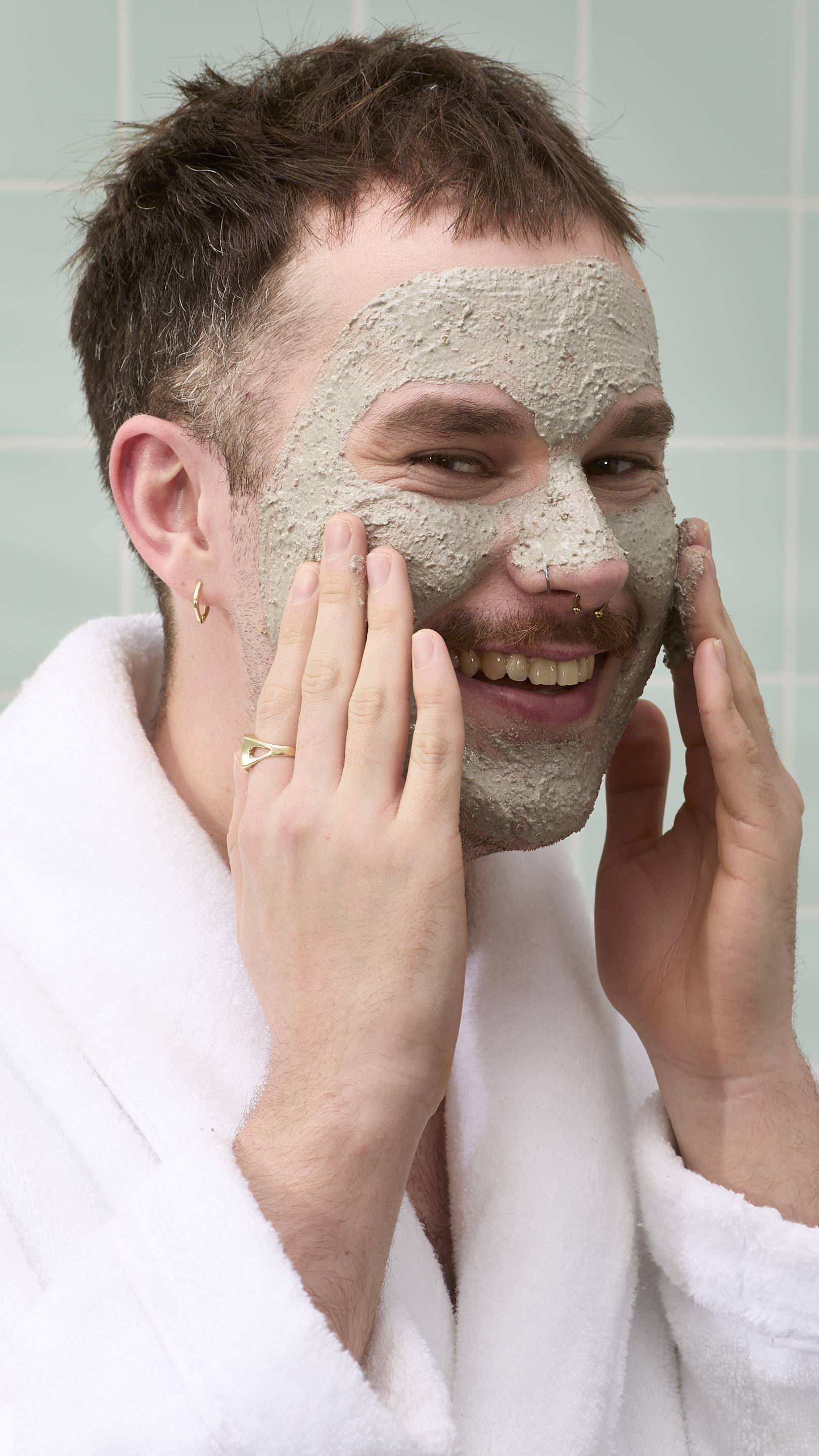 A close-up of a model with a moustache and nose piercings smiling while applying a textured grey "Movis" face mask with both hands. They are wearing a white dressing gown set against a background of light green bathroom tiles.