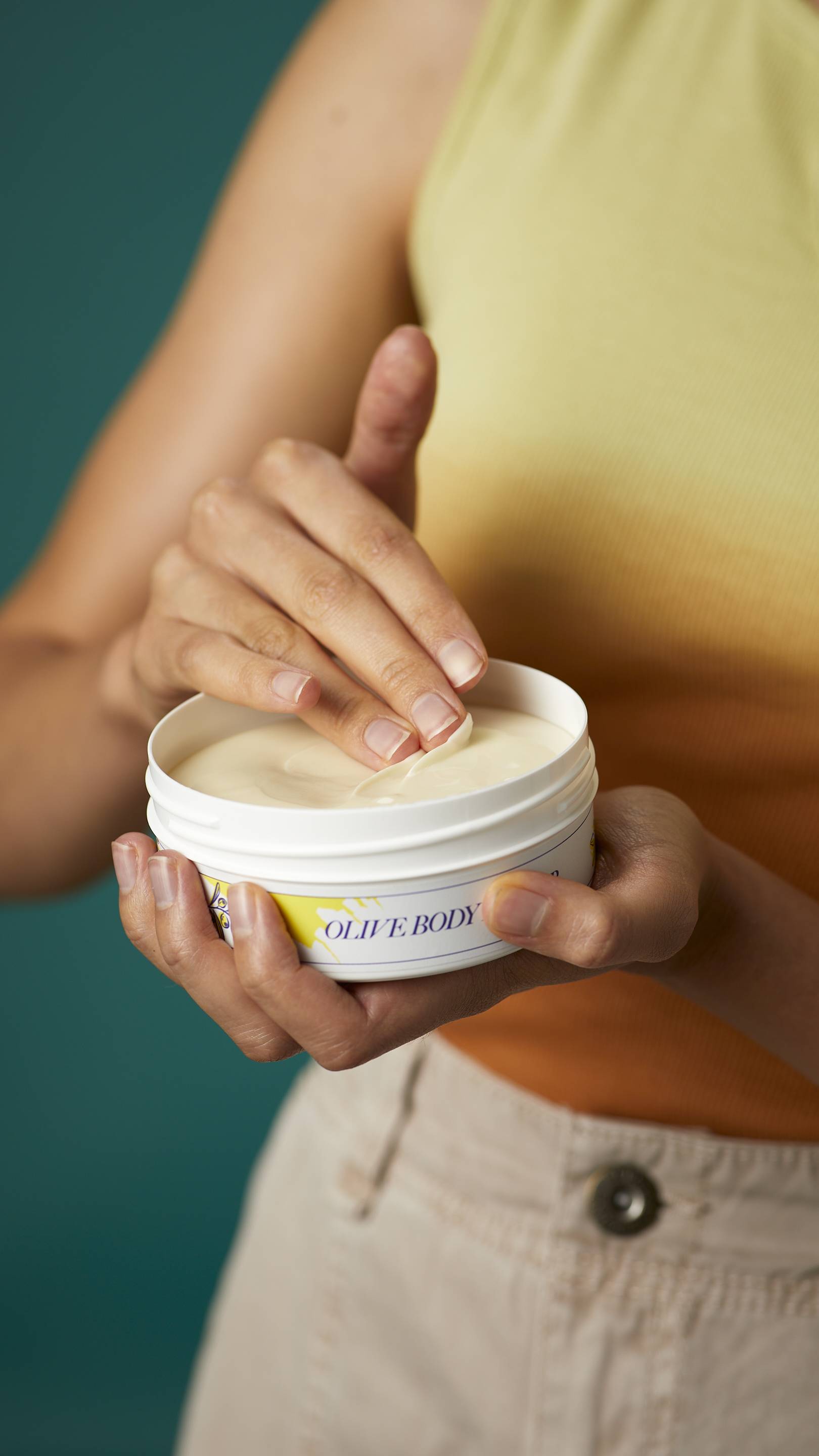 A close-up shot of the model's hands. One hand is holding the flat round, white, LUSH Version-themed packed tub of Olive body butter.