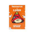 An enamel pin badge from the Paddington x Lush collection. The badge features Paddington Bear’s head wearing a red hat with a small yellow duck on top. It is displayed on an orange backing card with white bubbles and orange slice illustrations.