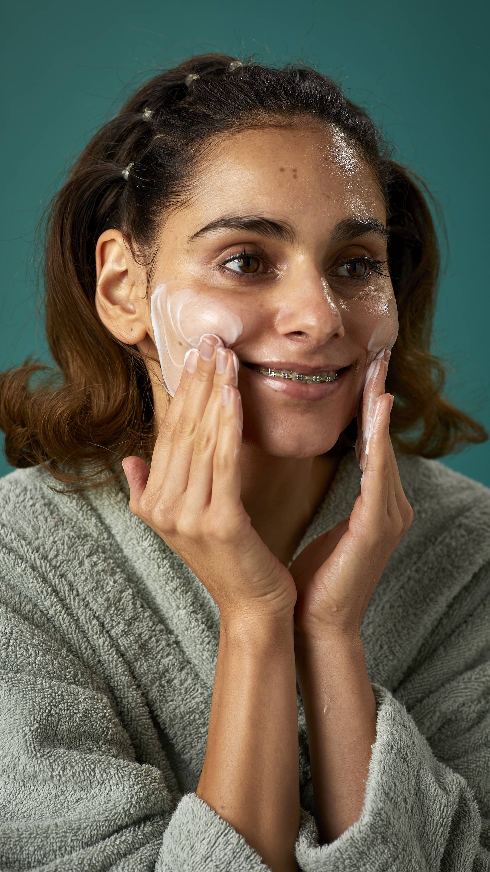 The image shows the model wrapped in a thick, sage-green dressing gown on a teal background. They are smiling, showing a glimpse of braces as they apply the Pineapple facial wash over their cheeks.