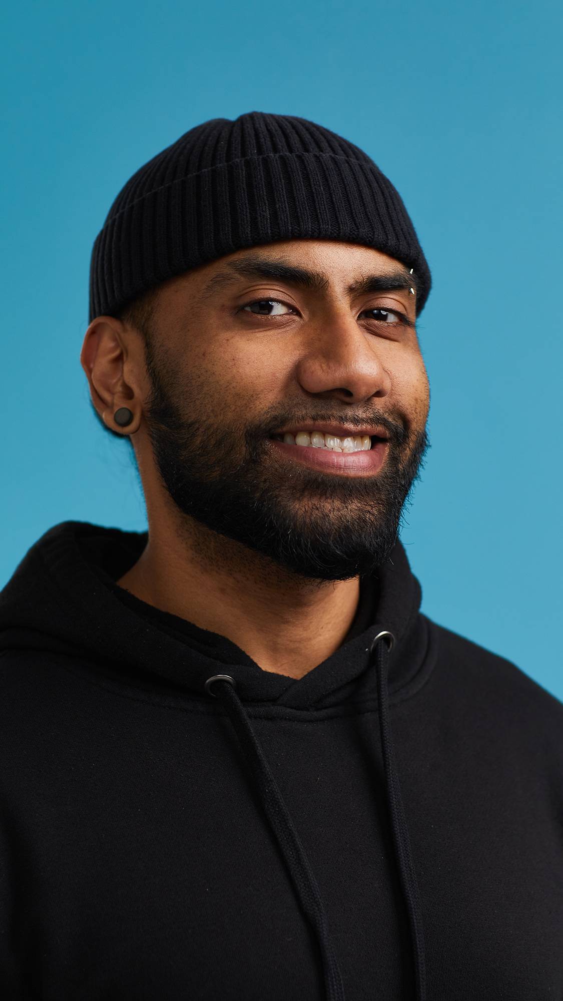 The image shows the model on a sky blue background wearing a back hoodie and hat. They have just brushed their teeth with the Plaque Sabbath toothpaste and are smiling at the camera with gleaming, shiny, fresh teeth.