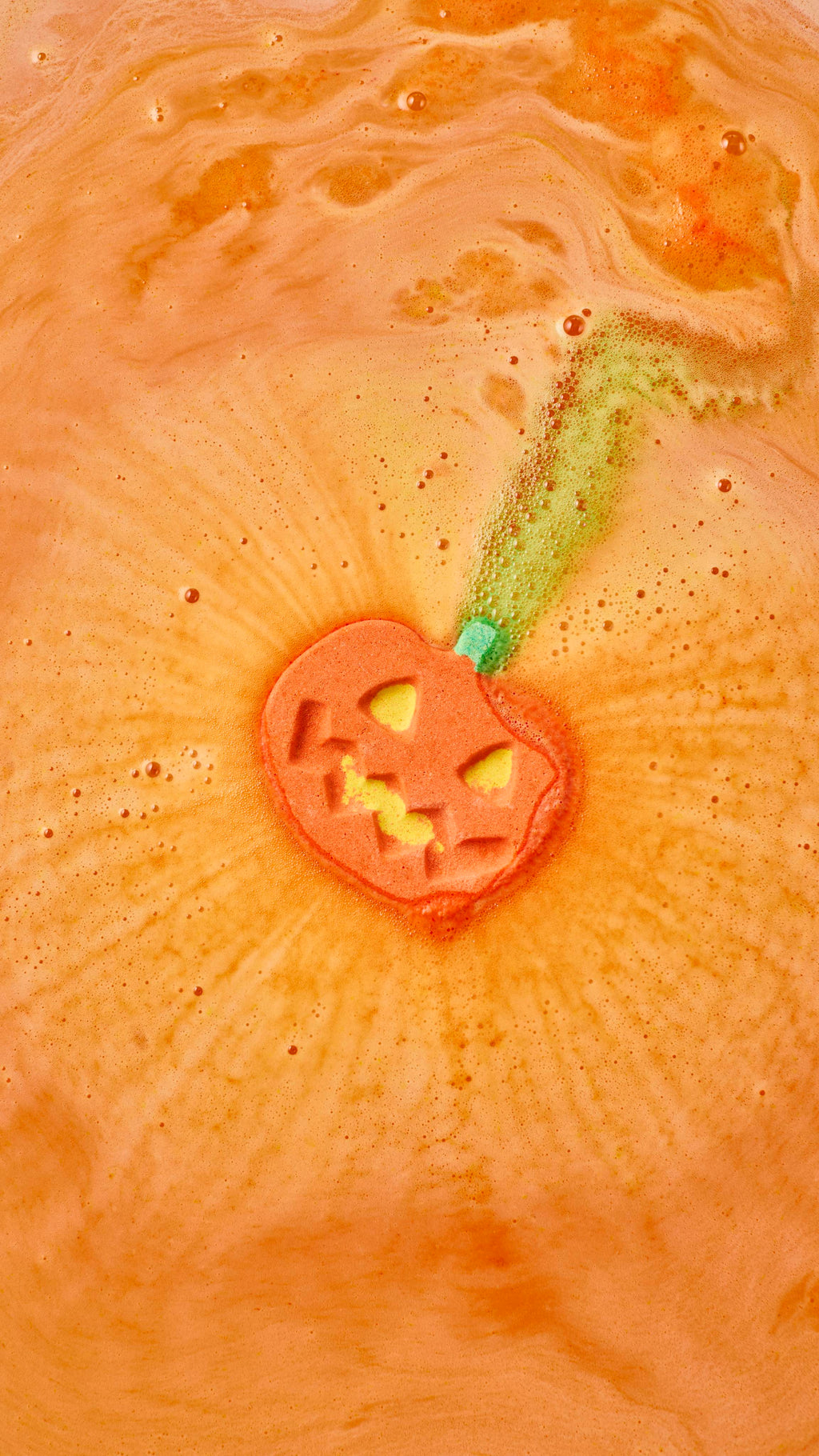 An orange pumpkin-shaped bath bomb dissolves in a bath, shooting a trail of bubbly green foam from its stalk. Concentric ripples of fine orange froth radiate outward from the carved jack-o'-lantern face into the warm orange water.