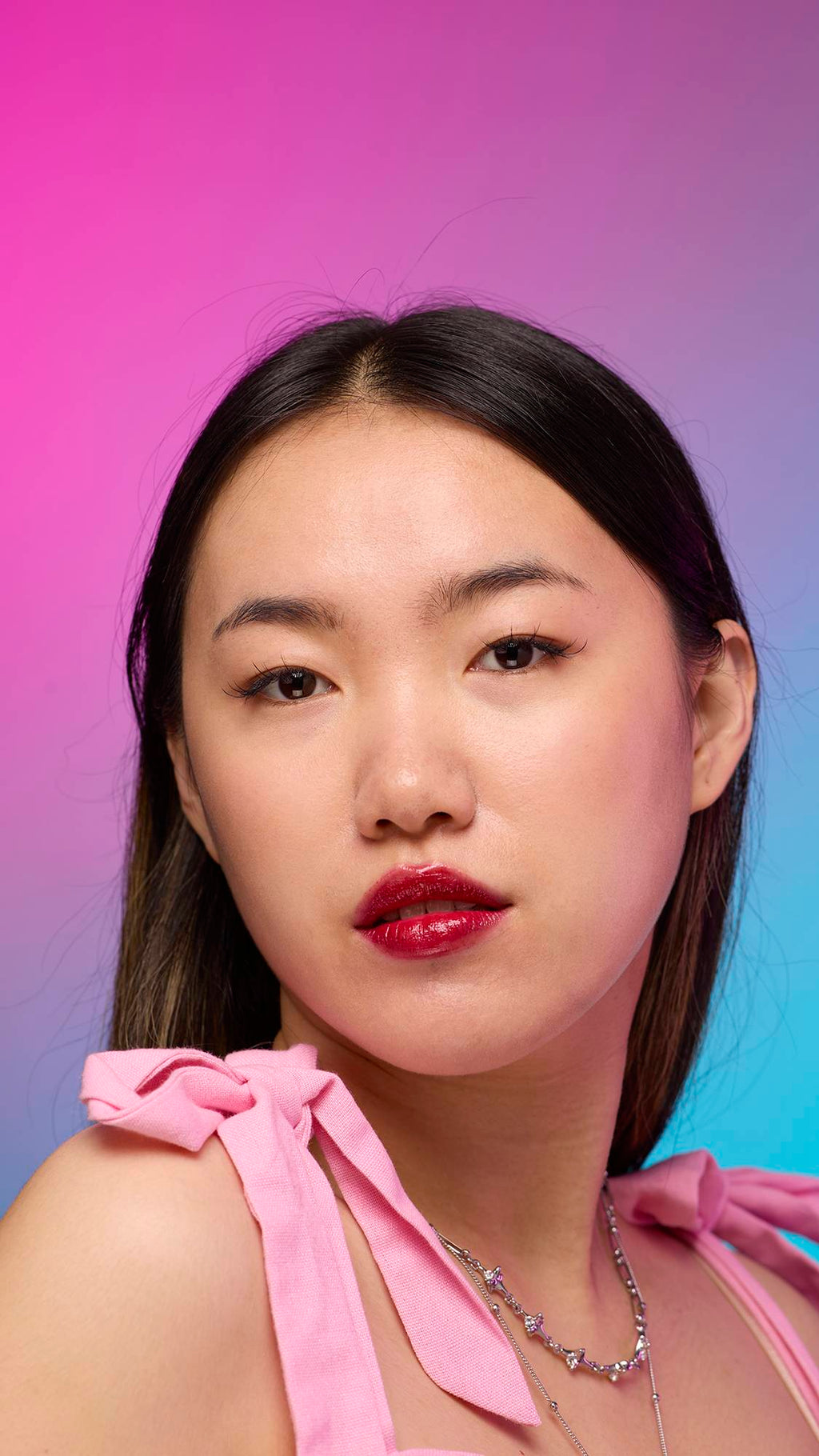 The model has long, straight dark hair and is on a pastel pink, blue and purple hue background. Their make-up has been done with the focal point being the juicy coating of carmine-red tint on their lips created by the Red Maple lip jelly.