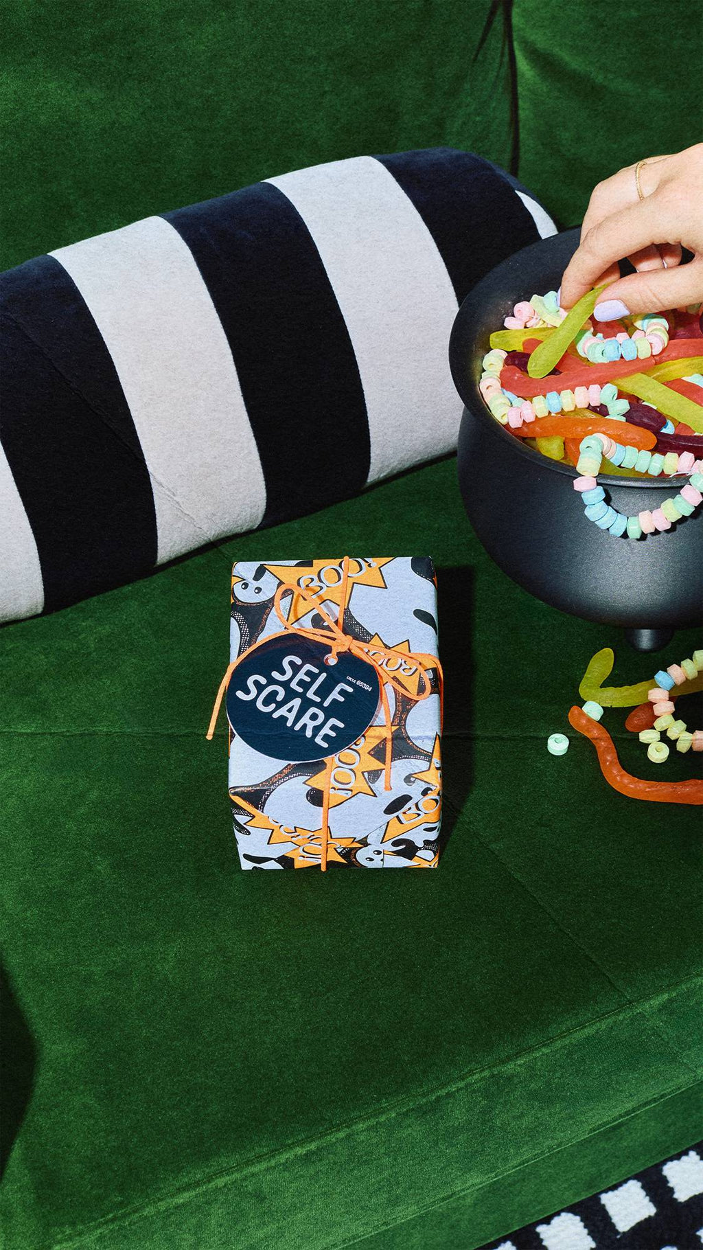A Lush 'Self Scare' wrapped gift box sits on a green velvet couch next to a black cauldron filled with colourful sweets. A hand reaches into the cauldron for a gummy worm, beside a black and grey striped cushion.
