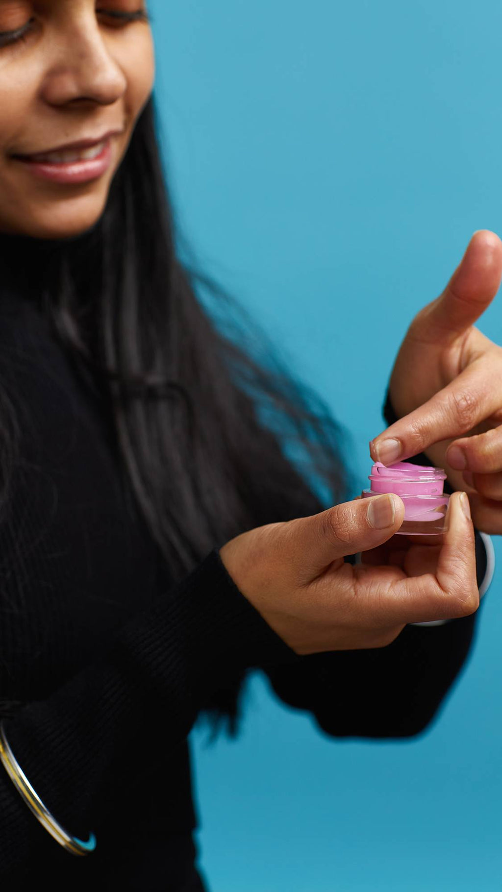 A close-up shot of the model's hands on a sky-blue background. They are holding a small, glass pot as they swipe the pretty, pink Sex Bomb lip gloss onto the tip of a finger.