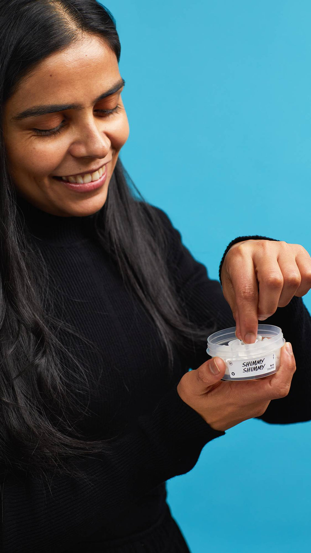 The image shows the model wearing a black turtleneck on a sky-blue background. They are holding a small, open clear, recycled plastic tub of Shimmy Shimmy mouthwash tabs and reaching in to pick one out.