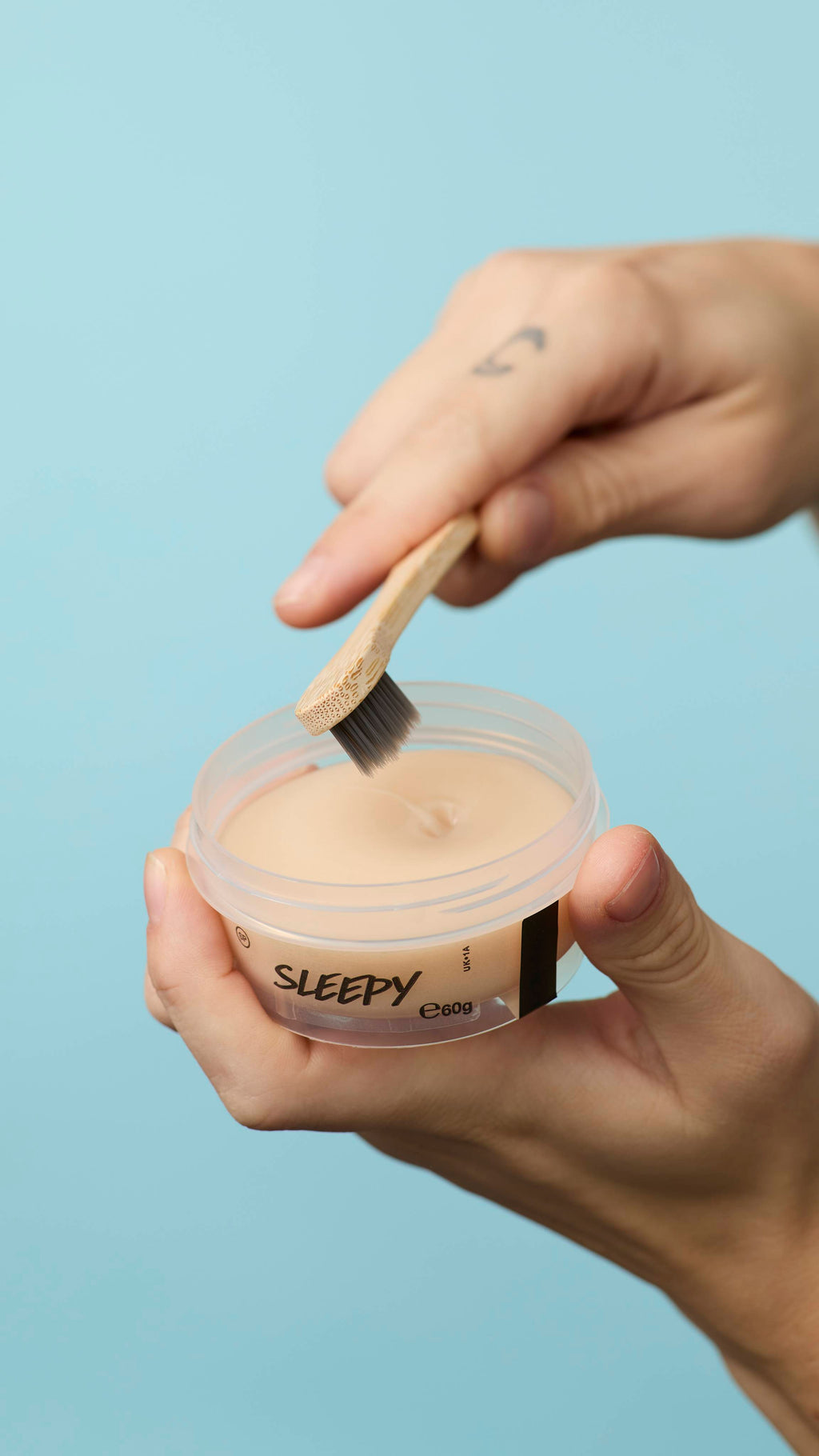 The image shows a pale blue background. The foreground shows a close-up of the model holding a clear, recycled plastic pot of LUSH Sleepy toothpaste with one hand as the other hand begins to dip the head of their toothbrush in.