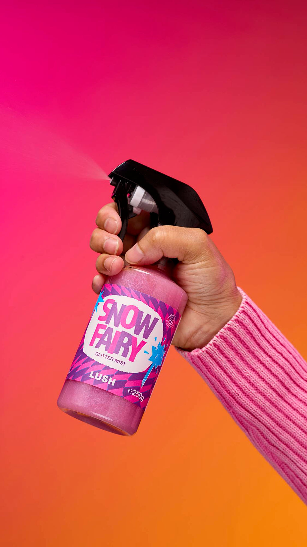 An arm is reaching from the right-hand side. The hand is spritzing a transparent, cylindrical plastic spray bottle with pink, shimmering liquid and a black top. A colourful label ready "Snow Fair Glitter Mist" on a pink-to-orange gradient background.