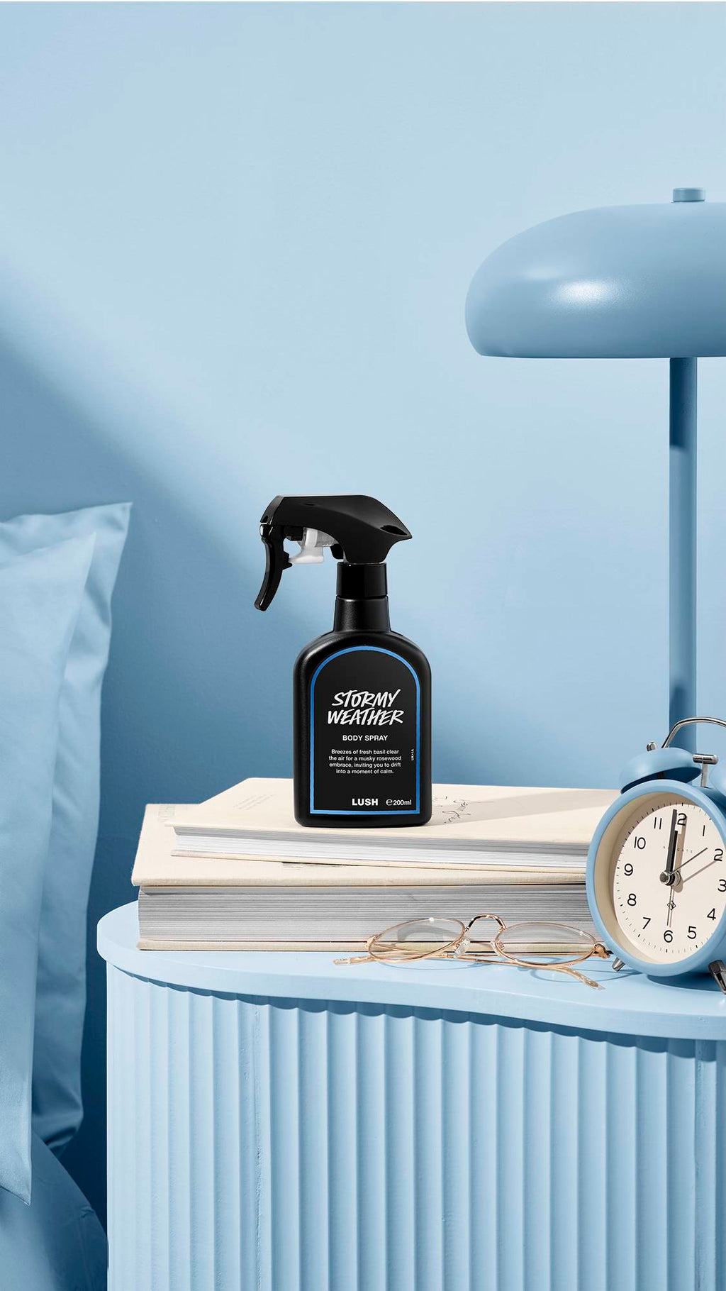 The image shows a recycled black plastic LUSH spray bottle of "Stormy Weather" sitting on a pile of books on a sea-blue bedside table. A matching lamp, alarm clock, and glasses sit nearby, next to a blue bed and wall.