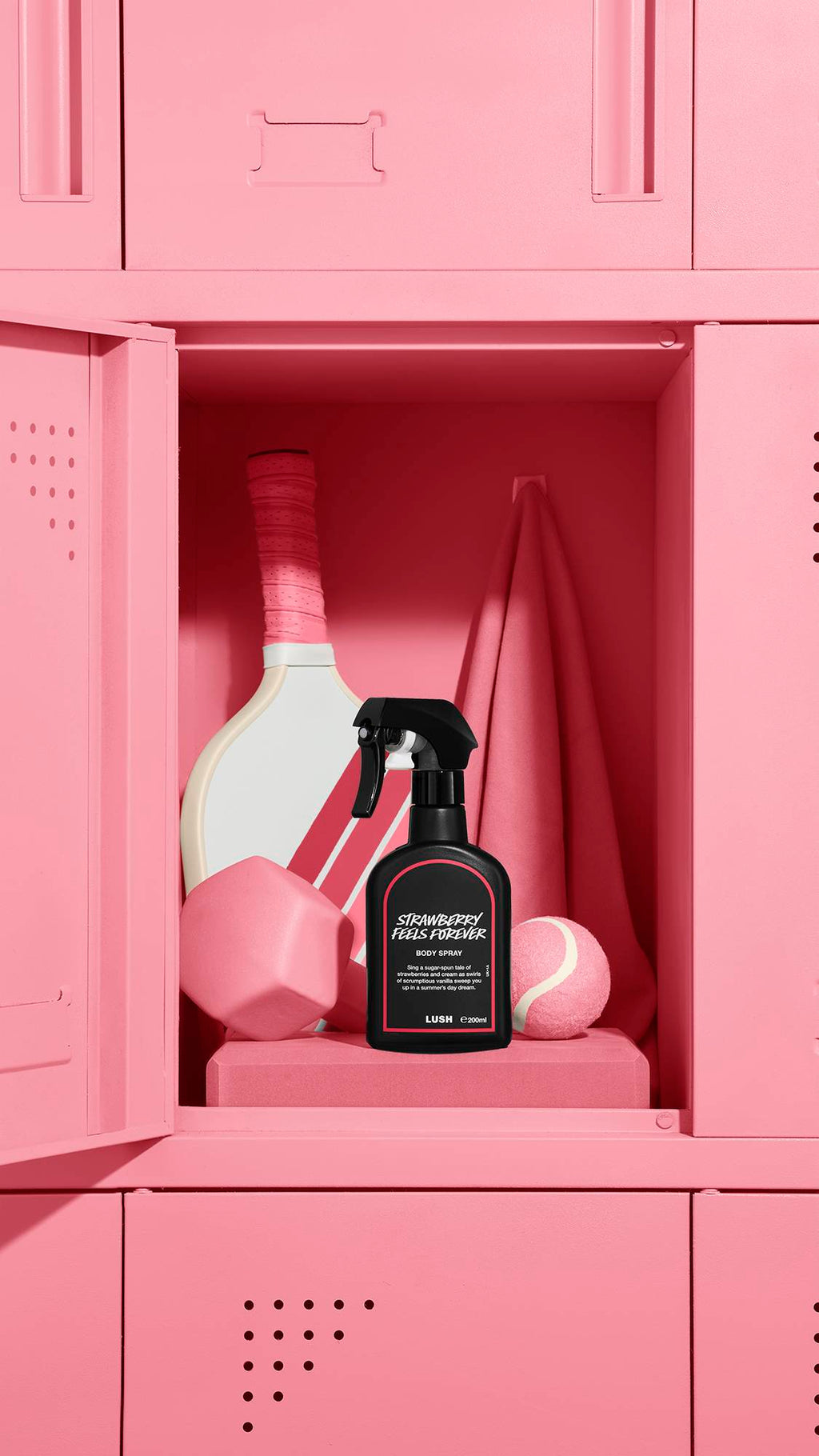 The image shows a black LUSH 'Strawberry Feels Forever' body spray bottle with a red rimmed label, standing inside an open pastel pink locker. A tennis racket, a pink towel, weight, and tennis ball are also visible within the locker.