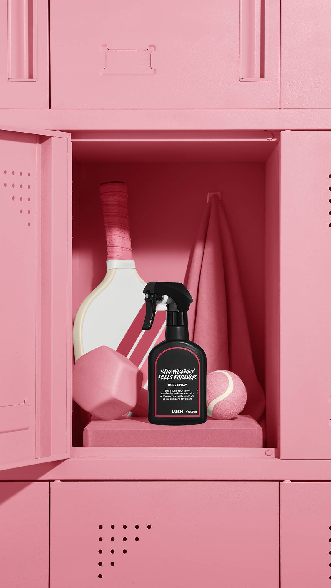 The image shows a black LUSH 'Strawberry Feels Forever' body spray bottle with a red rimmed label, standing inside an open pastel pink locker. A tennis racket, a pink towel, weight, and tennis ball are also visible within the locker.