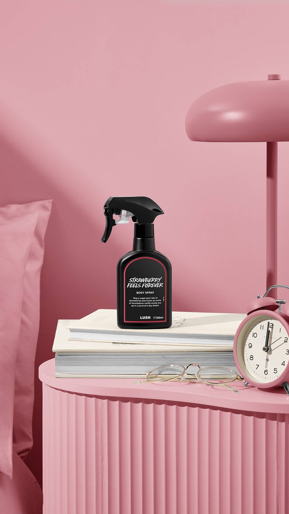 The image shows a recycled black plastic LUSH spray bottle of "Strawberry Feels Forever" sitting on a pile of books on a dusky pink bedside table. A matching coloured lamp, alarm clock, and glasses sit nearby, next to a pink bed and wall.