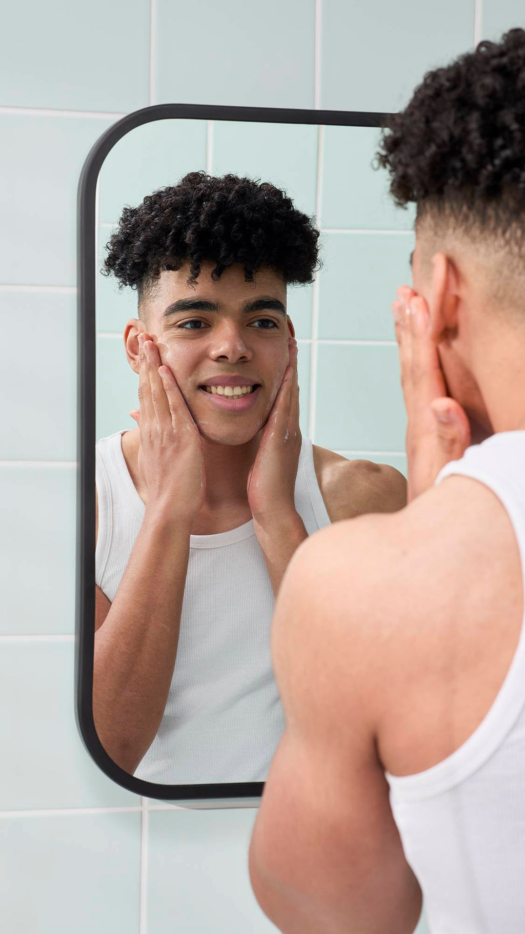 A reflection in a bathroom mirror shows a smiling model with short, curly dark hair applying a lather to their face, created by the Strawberry Picking facial gummy. They are wearing a white vest against a background of pale green wall tiles.