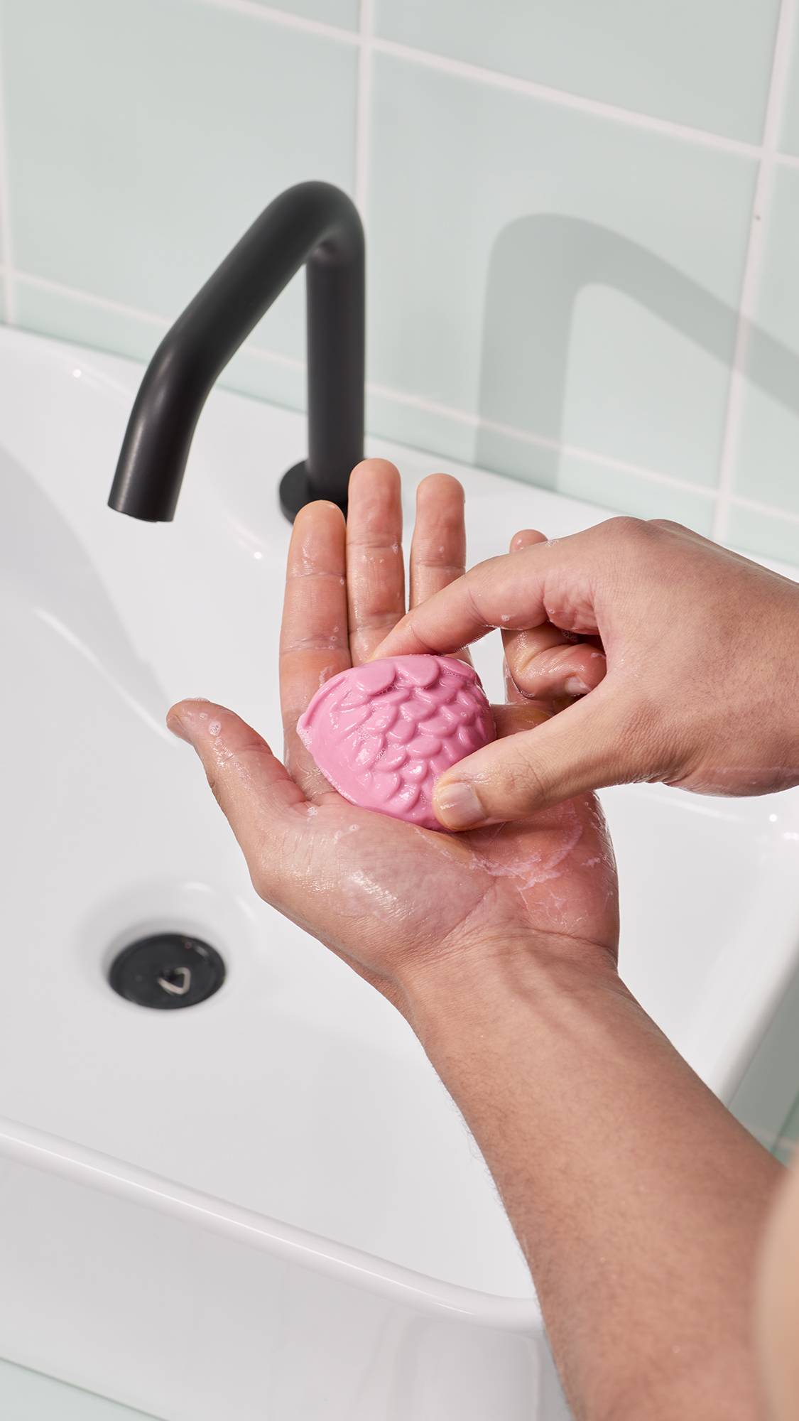 A model holds a pink, strawberry-shaped facial jelly in their hands to create a lather. The "Strawberry Picking" facial jelly cleanser has a scalloped texture. In the background, a white sink and black tap are visible against pale green wall tiles.