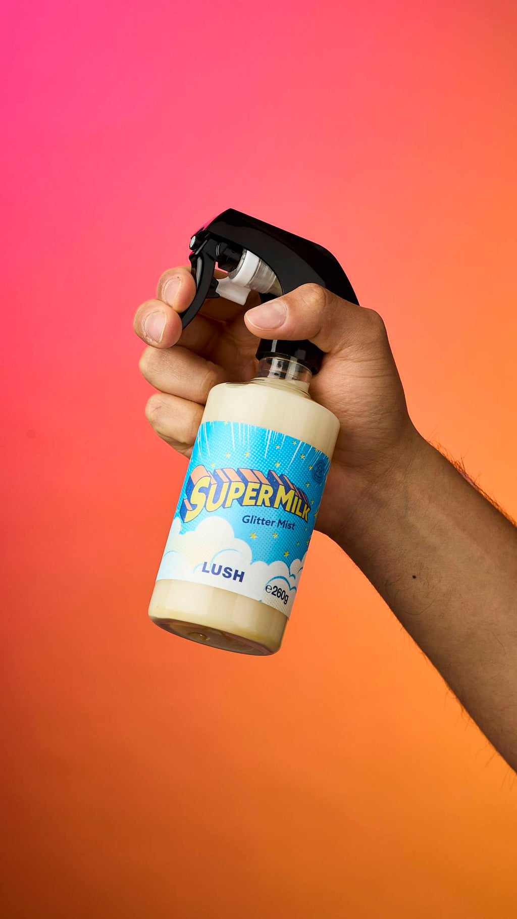 A hand holds a bottle of Lush 'Super Milk' Glitter Mist against a vibrant pink and orange gradient background. The bottle contains a cream-coloured liquid and has a bright blue, cloud-patterned label with bold yellow text.