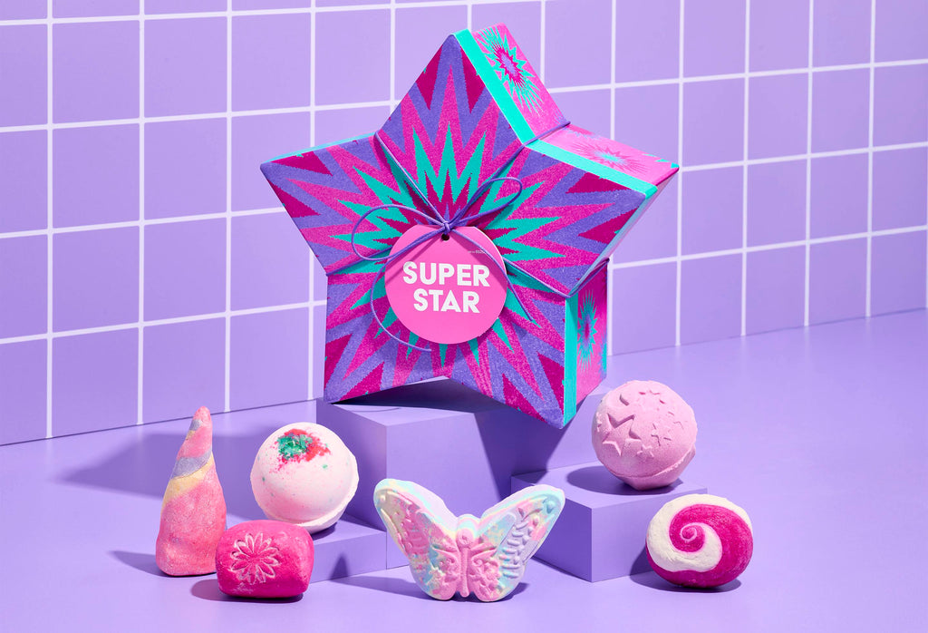 A star-shaped box with purple and turquoise zig-zags. Six colorful bath bombs and bubble bars. Displayed on purple blocks against a matching tiled background.