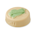Symbol of Love. A flat, circular, ivory-coloured solid balm with the word LUSH embossed on the side. On top is a pale green heart with a ribbon across which reads "MUM".