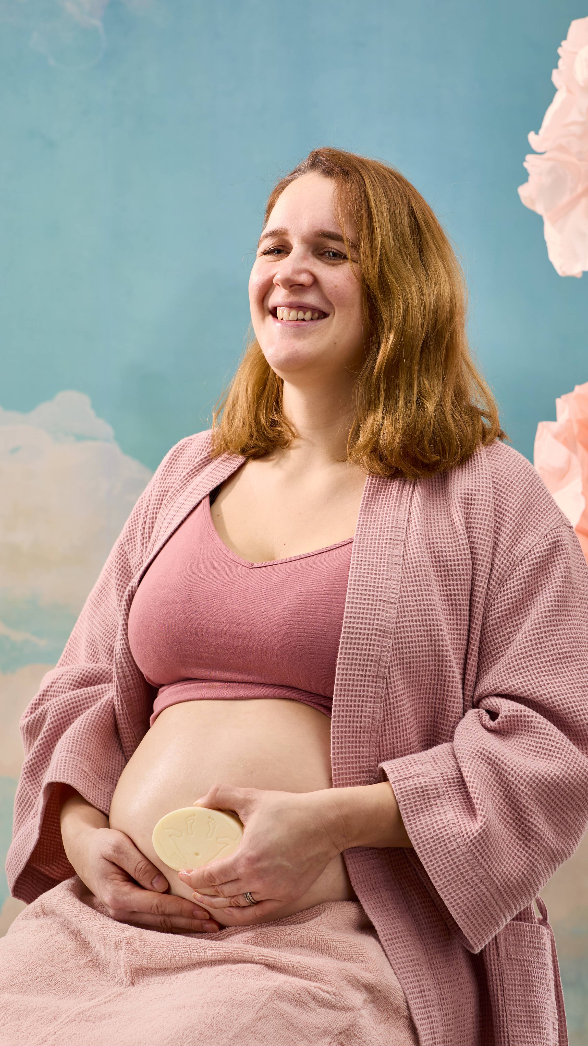 The image shows a pregnant model in a pastel pink cropped top, and dressing gown with a towel over their lap sitting on a blue sky background. They are holding their belly with one hand and smoothing The Bump massage bar on their skin.