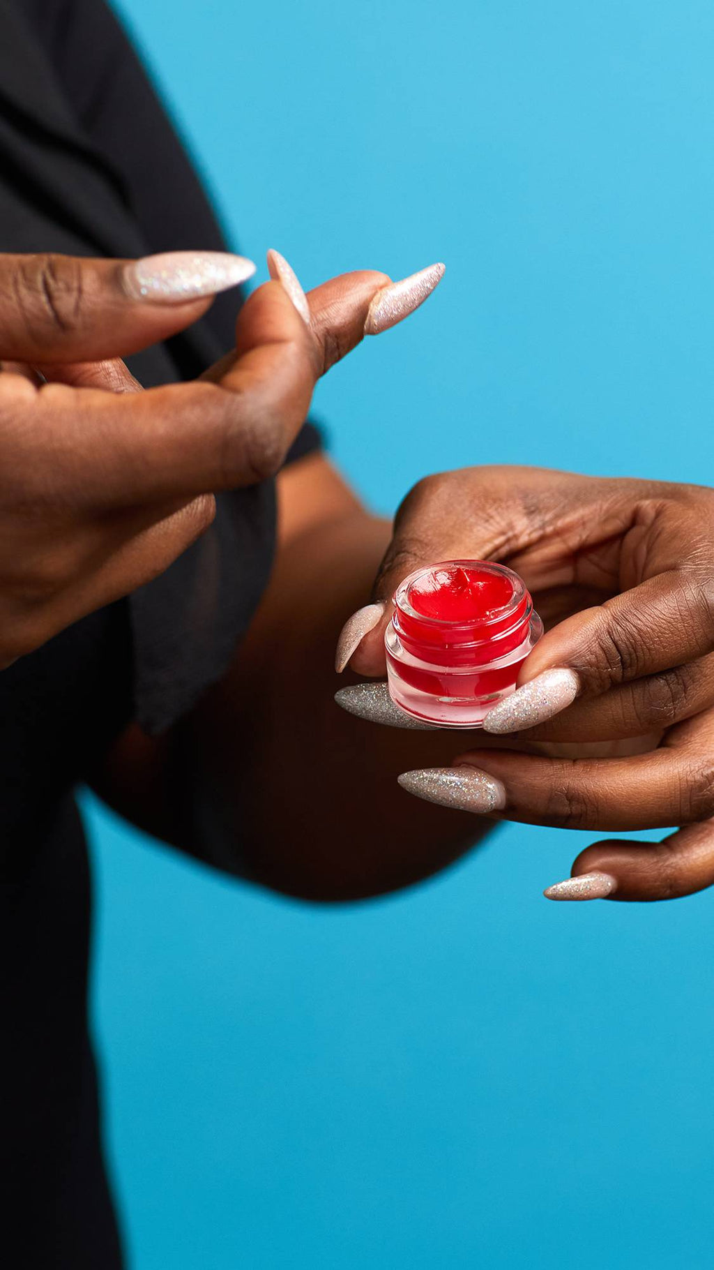 A close-up shot of the model's hands on a sky-blue background. They are holding a small, glass pot with sparkling, elegant nails as they swipe The Kiss lip gloss onto the tip of a finger.