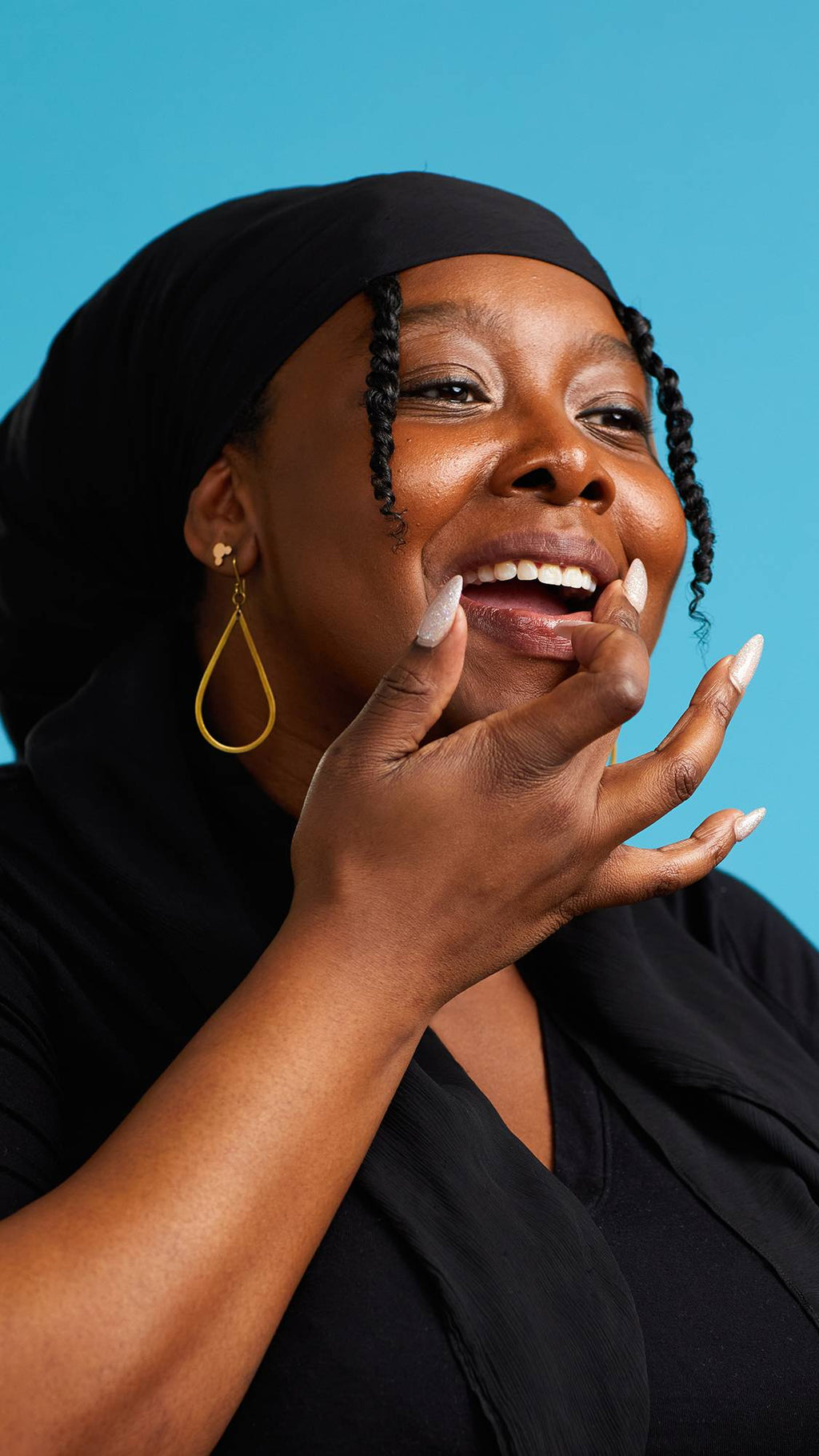 The image shows a close-up of the model on a sky-blue background. They are wearing a black top, black head scarf and gold earrings. Their mouth is parted as they apply The Kiss lip gloss to their lips.