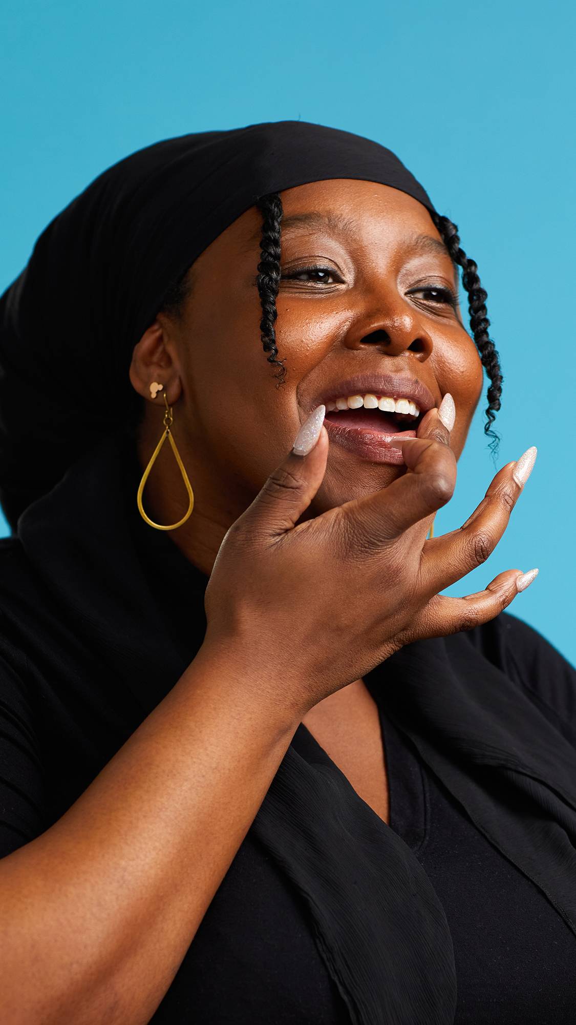 The image shows a close-up of the model on a sky-blue background. They are wearing a black top, black head scarf and gold earrings. Their mouth is parted as they apply The Kiss lip gloss to their lips.