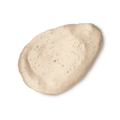 Tiny Musicians Of The Forest. A neatly smeared swatch of creamy, beige-coloured face mask with small dark exfoliating particles. The texture is rich and slightly lumpy, presented as a single dollop.