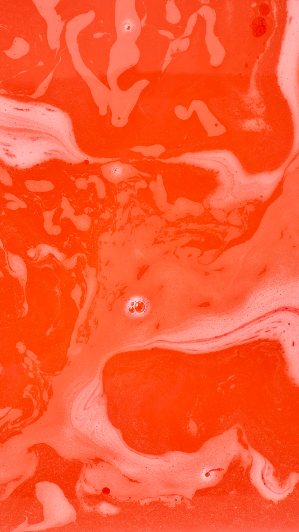 An abstract aerial shot of bath bomb foam swirling in water, creating marbled, wavy patterns of vibrant orange-red and pale pink-white froth. A few small clusters of bubbles are scattered across the colourful surface.