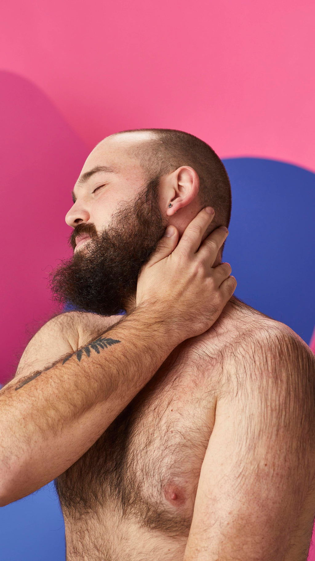 A shirtless, bearded person relaxes with eyes closed, gently massaging their neck with one hand to absorb the moisturizing oils from a Lush massage bar. Set against a vibrant abstract background of bold pink and blue shapes.