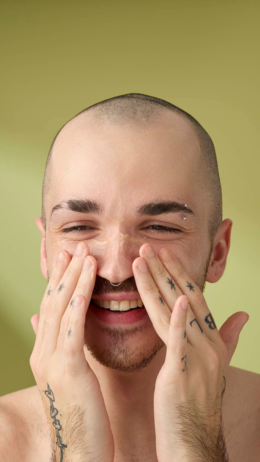 A close-up of a person with a shaved head and facial hair smiling as they massage a creamy 'Ultraplant' facial cleanser into their nose and cheeks, set against a soft olive-green background.