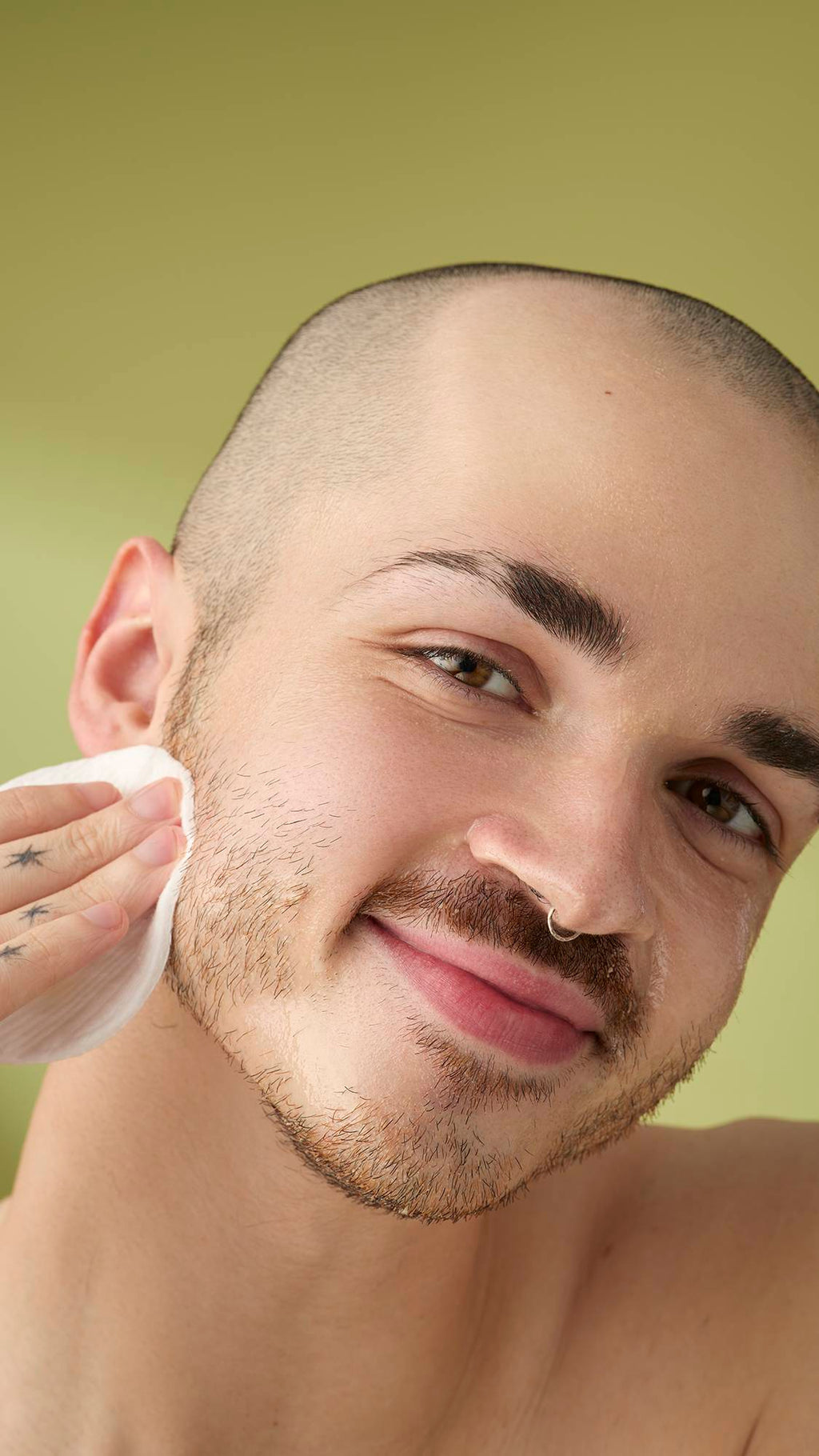 A close-up portrait of a person with a shaved head and facial hair, smiling as they wipe the 'Utlraplant' facial cleanser off their cheek with a white cotton pad, set against a soft olive-green background.