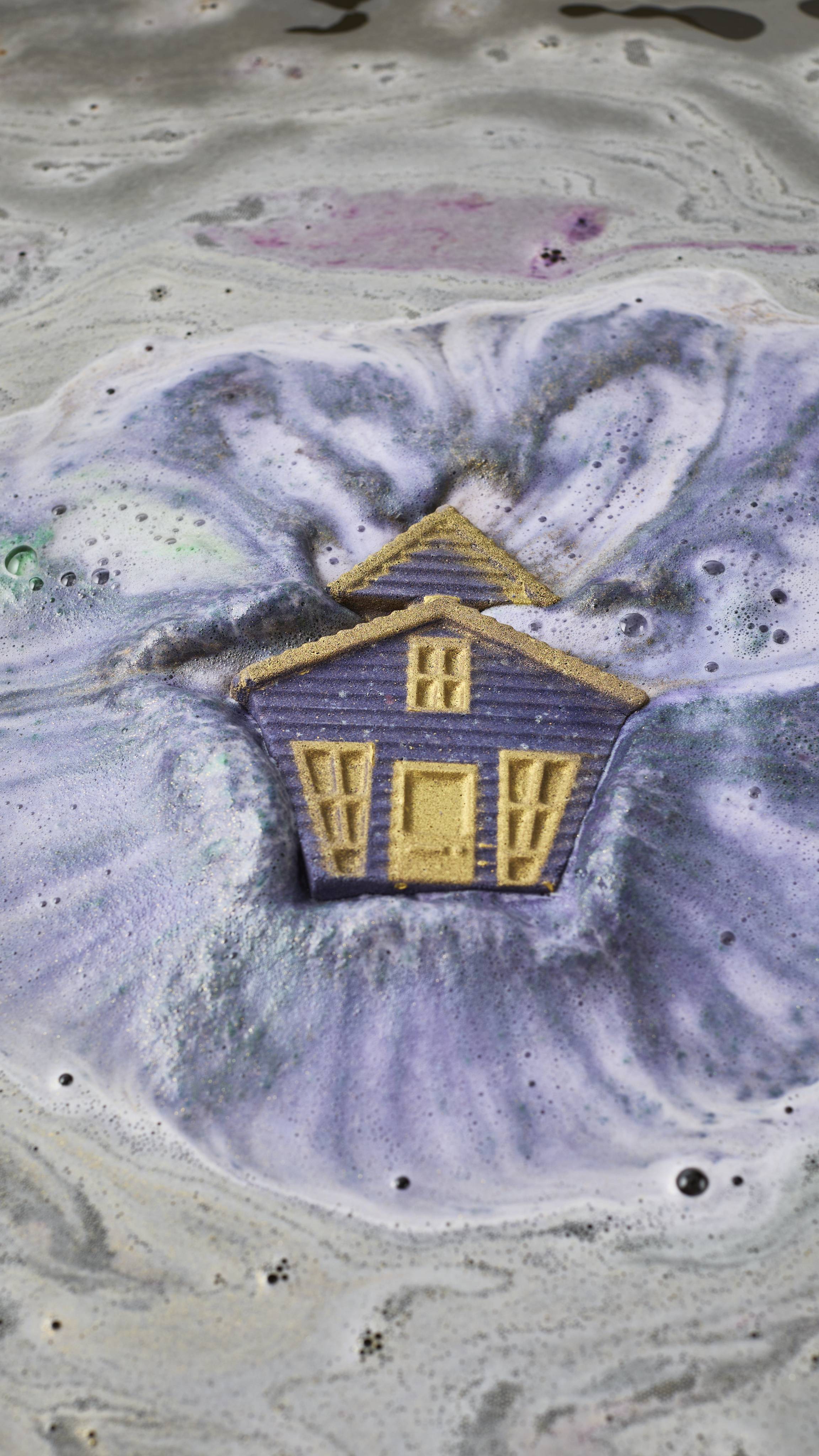 A purple haunted house bath bomb with gold windows and door fizzes and dissolves in murky bathwater, releasing thick swirls of purple and silver-grey foam and bubbles.